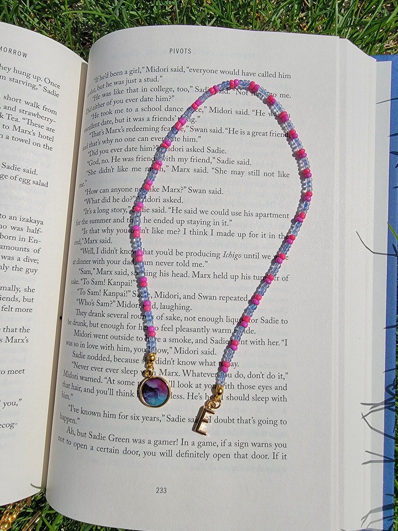 May include: A close-up of a book with a pink and blue beaded bookmark. The book is open to a page with text about a character named Marx. The text reads, "He was like that in college, too, Sadie. Not me."
