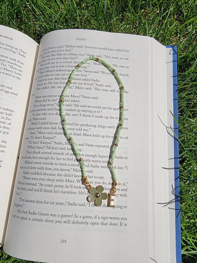 May include: A close-up of an open book with a green beaded necklace on top of it. The book is open to a page with the text "PIVOTS" at the top. The necklace has gold beads and a green flower charm.