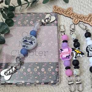 May include: A collection of beaded keychains with various charms, including bats, books, and a Kindle. The keychains feature a mix of pink, purple, black, white, and blue beads. One keychain is attached to a phone case with floral design.