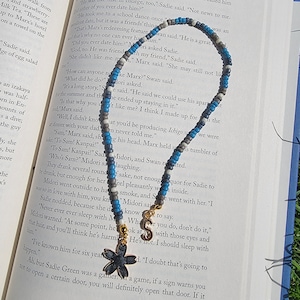 May include: A blue and grey beaded necklace with a gold charm in the shape of a flower. The necklace is lying on top of an open book with text about a character named Marx.