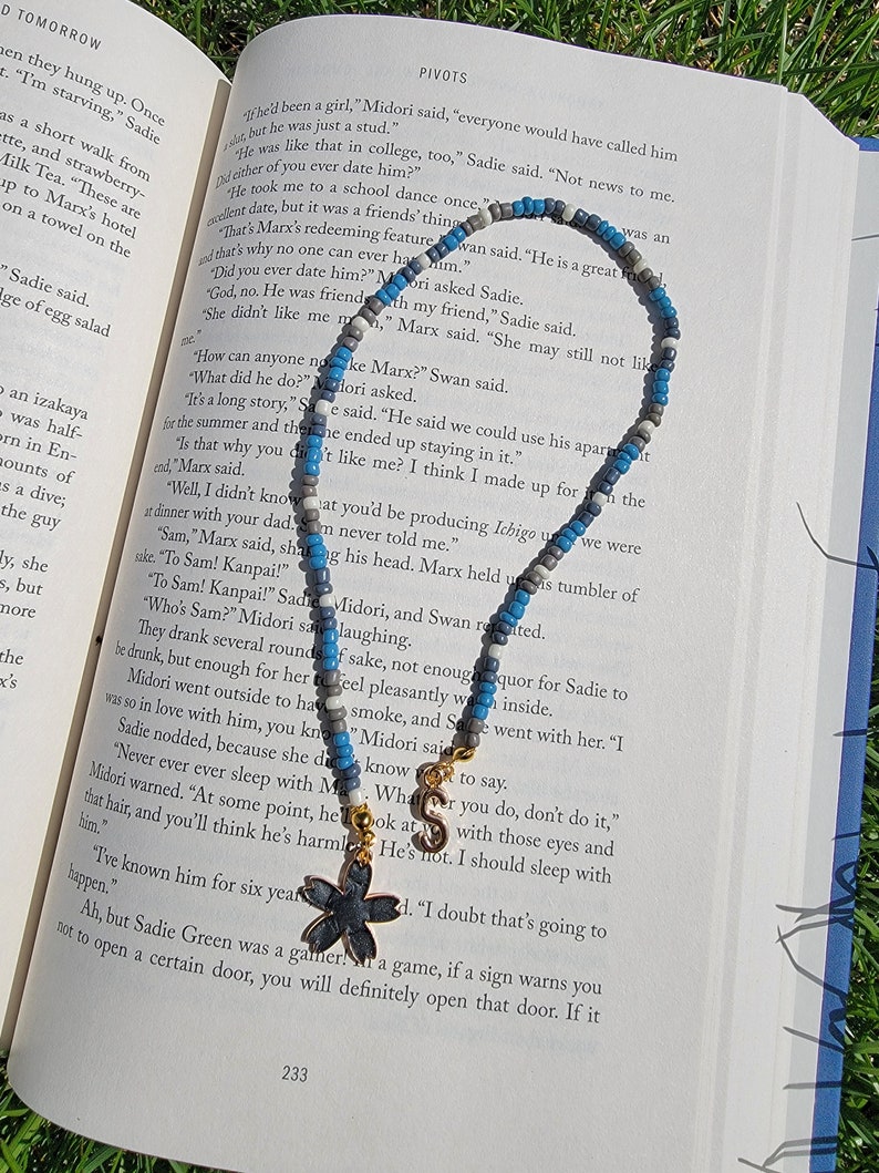 May include: A blue and grey beaded necklace with a gold charm in the shape of a flower. The necklace is lying on top of an open book with text about a character named Marx.