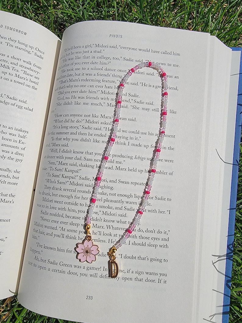 May include: A close-up of a book with a pink and white beaded necklace with a pink cherry blossom charm. The book is open to a page with the text "PIVOTS" and the text "She didn't like me much, "Marx said. "She may still like..."