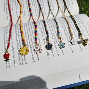May include: A set of colorful beaded bookmarks with various charms, including a red apple, a yellow daisy, a white daisy, a black flower, a blue flower, and a bee. The bookmarks are laid out on a book with text.