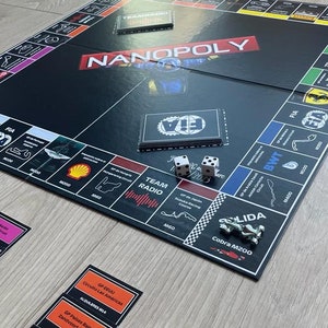 Puede incluir: Un juego de mesa Nanopoly negro con tarjetas de propiedad, dados y fichas. El tablero presenta circuitos de carreras y el texto "NANOPOLY" en rojo. El juego está sobre una superficie de madera clara.