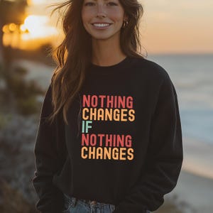 Puede incluir: Sudadera negra con un gráfico de estilo retro que dice "Nothing Changes If Nothing Changes".