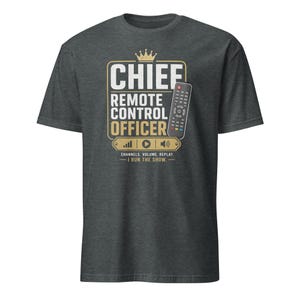 Può includere: Una t-shirt grigio scuro a maniche corte con una grafica che include un'icona a forma di corona, un telecomando e il testo CHIEF REMOTE CONTROL OFFICER, seguito da CHANNELS. VOLUME. REPLAY. — I RUN THE SHOW. — in caratteri bianchi e oro.