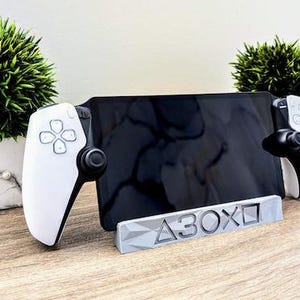 K&ouml;nnte beinhalten: Ein wei&szlig;-schwarzer Gaming-Controller-St&auml;nder mit einer grauen Basis, auf der "A3OX" steht. Der St&auml;nder h&auml;lt ein schwarzes Tablet und zwei wei&szlig;e Gaming-Controller.