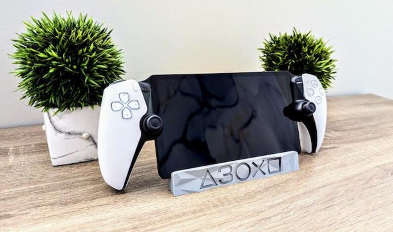 K&ouml;nnte beinhalten: Ein wei&szlig;-schwarzer Gaming-Controller-St&auml;nder mit einer grauen Basis, auf der "A3OX" steht. Der St&auml;nder h&auml;lt ein schwarzes Tablet und zwei wei&szlig;e Gaming-Controller.