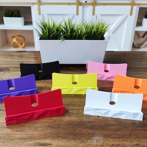 K&ouml;nnte beinhalten: Sieben Telefonst&auml;nder in verschiedenen Farben, darunter Lila, Rot, Gelb, Wei&szlig;, Orange, Rosa und Schwarz. Die St&auml;nder sind aus einem strukturierten Material gefertigt und haben ein Loch in der Mitte f&uuml;r Ladekabel.