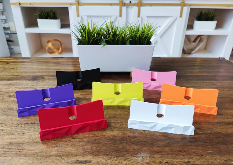 K&ouml;nnte beinhalten: Sieben Telefonst&auml;nder in verschiedenen Farben, darunter Lila, Rot, Gelb, Wei&szlig;, Orange, Rosa und Schwarz. Die St&auml;nder sind aus einem strukturierten Material gefertigt und haben ein Loch in der Mitte f&uuml;r Ladekabel.
