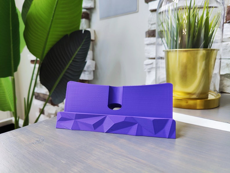 K&ouml;nnte beinhalten: Ein lilafarbener Telefonst&auml;nder mit geometrischem Design. Der St&auml;nder ist aus Kunststoff und hat einen Schlitz zum Halten eines Telefons.