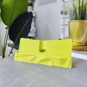 K&ouml;nnte beinhalten: Ein leuchtend gelber Telefonst&auml;nder mit geometrischem Design. Der St&auml;nder ist aus Kunststoff und hat einen Schlitz zum Halten eines Telefons.