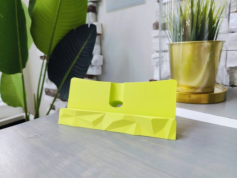 K&ouml;nnte beinhalten: Ein leuchtend gelber Telefonst&auml;nder mit geometrischem Design. Der St&auml;nder ist aus Kunststoff und hat einen Schlitz zum Halten eines Telefons.