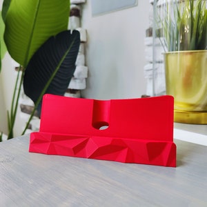 K&ouml;nnte beinhalten: Ein roter Telefonst&auml;nder mit geometrischem Design. Der St&auml;nder ist aus einem glatten, kunststoffartigen Material gefertigt.