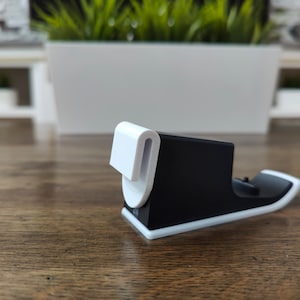 May include: A black and white phone stand with a white curved hook on the top. The stand is made of plastic and has a smooth finish.