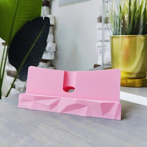 K&ouml;nnte beinhalten: Ein rosa Telefonst&auml;nder aus Kunststoff mit geometrischem Design. Der St&auml;nder hat einen Ausschnitt auf der R&uuml;ckseite f&uuml;r Ladekabel.