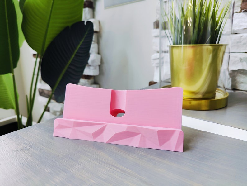 K&ouml;nnte beinhalten: Ein rosa Telefonst&auml;nder aus Kunststoff mit geometrischem Design. Der St&auml;nder hat einen Ausschnitt auf der R&uuml;ckseite f&uuml;r Ladekabel.