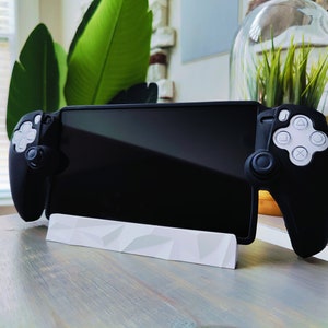 May include: A black gaming controller with white buttons and a black phone or tablet attached to it. The controller is sitting on a white stand on a wooden table.