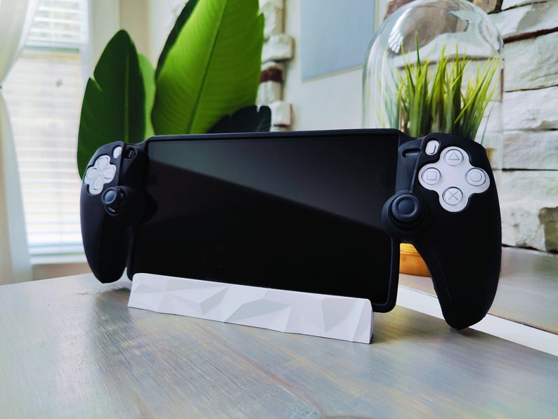 K&ouml;nnte beinhalten: Ein schwarzer Gamecontroller mit wei&szlig;en Kn&ouml;pfen und einem schwarzen Telefon oder Tablet, das daran befestigt ist. Der Controller steht auf einem wei&szlig;en St&auml;nder auf einem Holztisch.