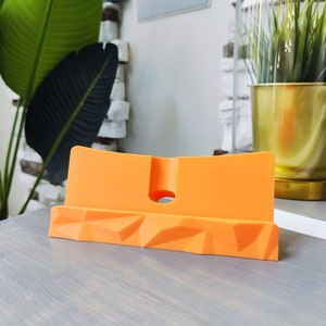 K&ouml;nnte beinhalten: Orangener Telefonst&auml;nder mit geometrischem Design. Der St&auml;nder ist aus Kunststoff und hat ein kleines Loch auf der R&uuml;ckseite f&uuml;r Ladekabel.