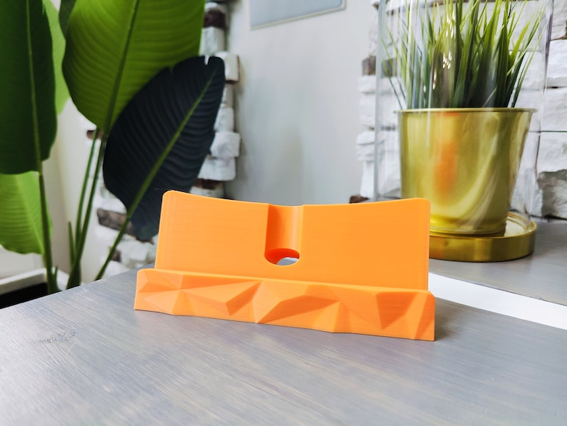 K&ouml;nnte beinhalten: Orangener Telefonst&auml;nder mit geometrischem Design. Der St&auml;nder ist aus Kunststoff und hat ein kleines Loch auf der R&uuml;ckseite f&uuml;r Ladekabel.