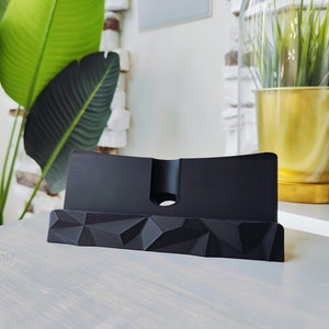 K&ouml;nnte beinhalten: Ein schwarzer Telefonst&auml;nder mit geometrischem Design. Der St&auml;nder ist aus hartem Kunststoff gefertigt und hat einen erh&ouml;hten Rand, damit Ihr Telefon nicht verrutscht.
