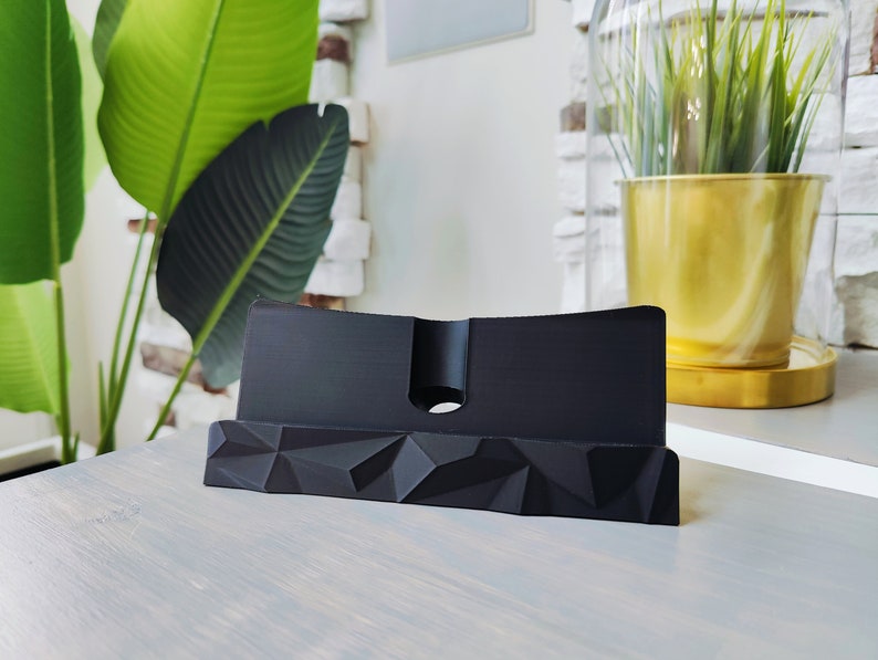 K&ouml;nnte beinhalten: Ein schwarzer Telefonst&auml;nder mit geometrischem Design. Der St&auml;nder ist aus hartem Kunststoff gefertigt und hat einen erh&ouml;hten Rand, damit Ihr Telefon nicht verrutscht.