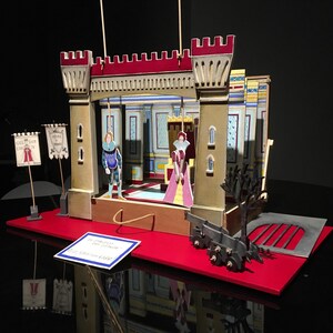 May include: A miniature stage set with a red and gold castle backdrop, featuring two puppets, a king and a queen, on a red stage. The stage is set on a black surface with a red base.