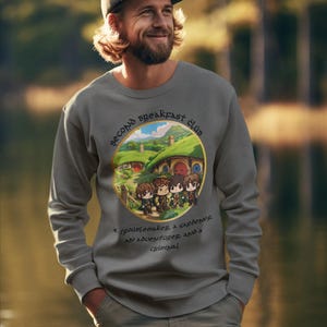 Op de afbeelding: Een grijs sweatshirt met een cirkelvormige afbeelding van een hobbitdorp en de tekst "Second Breakfast Club" in een gouden cirkel. De tekst onder de cirkel luidt "A groundskeeper, a gardener, an adventurer, and a criminal."