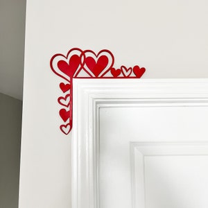 May include: Red paper hearts of various sizes are arranged in a corner design on a white wall, above a white door frame.