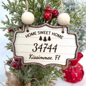 Op de afbeelding: Houten kerstversiering met een witte achtergrond en een bruine rand. De versiering heeft de tekst "Home Sweet Home" met een rij dennenbomen en het nummer "34744". De tekst "Kissimmee, FL" is ook op de versiering te vinden.