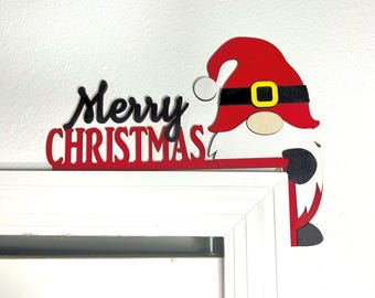 May include: A wooden Christmas decoration featuring a gnome wearing a red hat with a black belt and yellow buckle. The words "Merry Christmas" are displayed in black and red, perfect for holiday decor.
