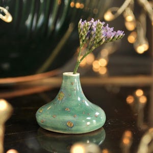 Op de afbeelding: Een kleine, handgemaakte keramische vaas in een turquoise groene tint, bespikkeld met bruine en gouden accenten. De vaas bevat een takje paarse bloemen. De vaas heeft een bolvormige basis en een smalle hals. Decoratieve lichten zijn op de achtergrond.