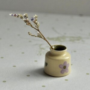 Op de afbeelding: Een kleine, crèmekleurige keramische vaas met een paars bloemmotief en een takje gedroogde bloemen. De vaas heeft een donkergroen interieur en staat op een gestippelde ondergrond. Een delicaat bloemstuk.