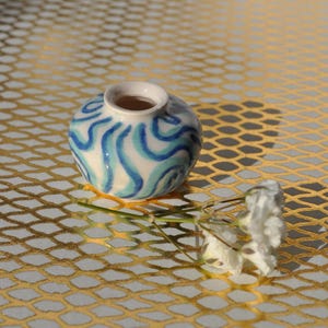 Pode incluir: Um pequeno vaso de cerâmica redondo, com base branca e padrão de ondas azuis. O vaso tem uma abertura estreita e está sobre uma superfície dourada de malha. Um raminho de flores brancas está por perto.
