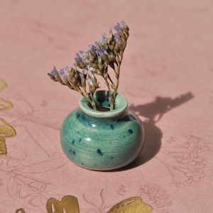 Op de afbeelding: Een kleine, ronde, turquoise keramische vaas met blauwe spikkels bevat gedroogde paarse bloemen. De vaas staat op een roze oppervlak met bloemenpatroon, met gouden accenten.