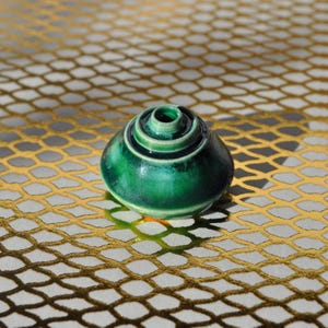 May include: A small, green ceramic vase with a spiral design. The vase has a glossy finish and a small opening at the top. The vase is sitting on a gold-coloured, patterned surface. The vase is a decorative item.