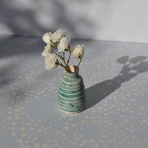Op de afbeelding: Een kleine, turquoise keramische vaas met horizontale witte strepen en een beige binnenkant, met witte bloemen. De vaas staat op een lichtblauwe ondergrond met een stippenpatroon en werpt een schaduw.