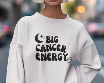May include: White sweatshirt with the text "BIG CANCER ENERGY" in black, featuring a crescent moon, stars, and the Cancer zodiac symbol. The sweatshirt is worn by a person with short, blonde hair.