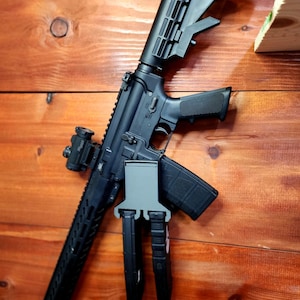 May include: Black rifle with a red dot sight and a magazine attached. The rifle is mounted on a wooden surface. The rifle has a black stock and grip. The magazine is also black.