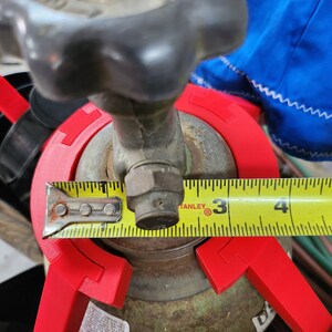 May include: A yellow measuring tape is wrapped around a rusty metal tank with a red plastic safety device. The tape reads 3 inches. The tank has a label that says "LOT NUMBER C190 G 02 045 RJ".