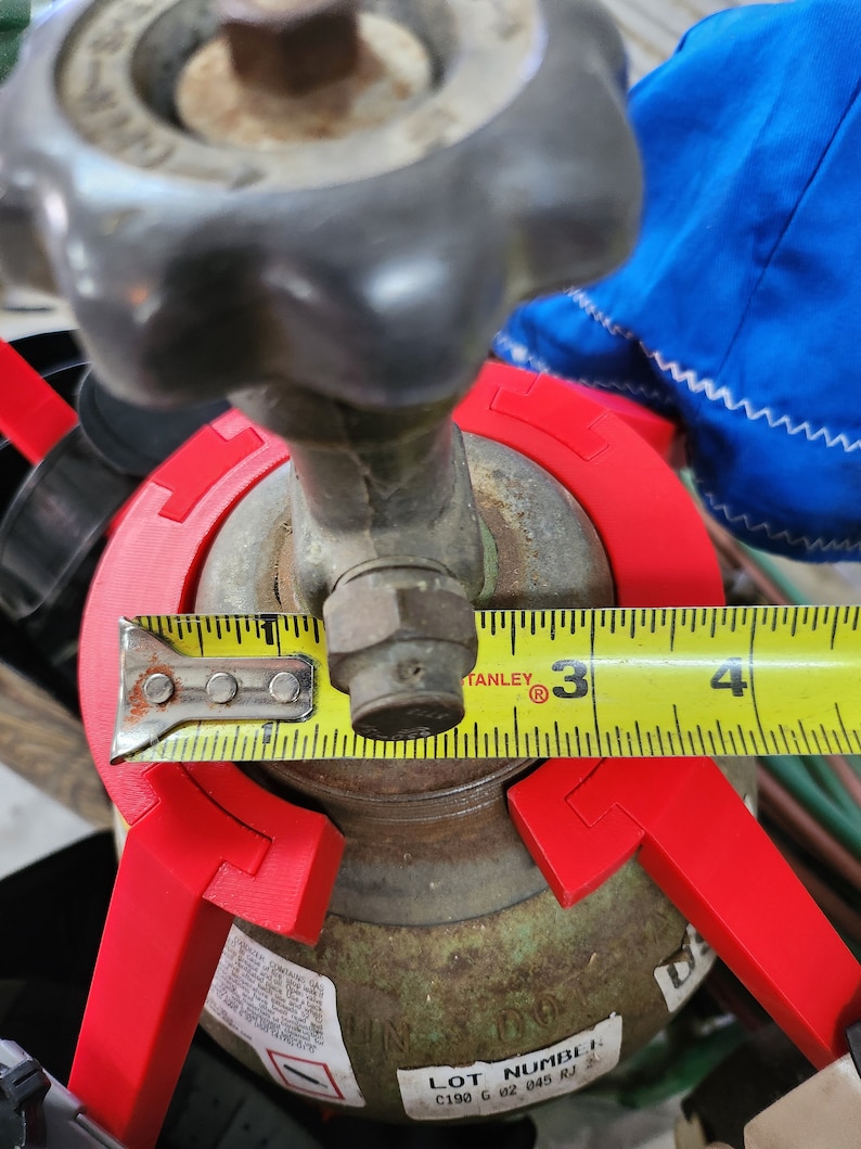 May include: A yellow measuring tape is wrapped around a rusty metal tank with a red plastic safety device. The tape reads 3 inches. The tank has a label that says "LOT NUMBER C190 G 02 045 RJ".