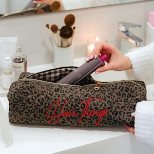 May include: A brown and black leopard print quilted cosmetic bag with the words "Hair Things" embroidered in red. A person is holding a gray and pink hair styling tool. The bag is open, revealing a brown and white checkered lining.