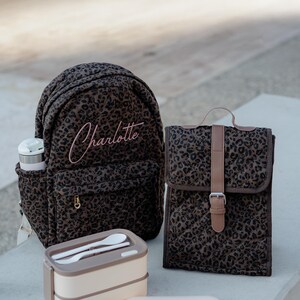 K&ouml;nnte beinhalten: Ein Set aus braunen Accessoires mit Leopardenmuster, darunter ein Rucksack, eine Lunchtasche und eine Lunchbox. Der Rucksack ist mit dem Namen "Charlotte" in Rosa bestickt. Die Lunchbox enth&auml;lt Utensilien.