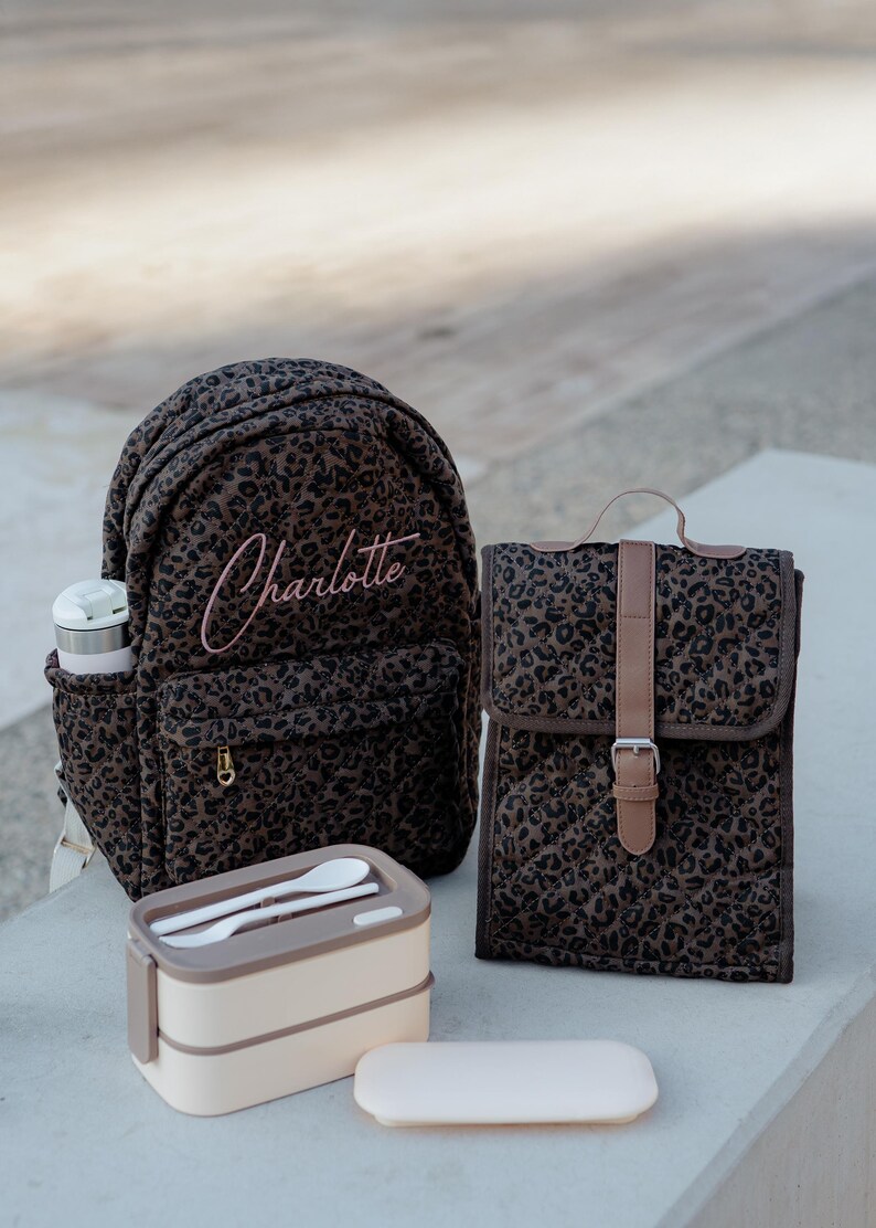 K&ouml;nnte beinhalten: Ein Set aus braunen Accessoires mit Leopardenmuster, darunter ein Rucksack, eine Lunchtasche und eine Lunchbox. Der Rucksack ist mit dem Namen "Charlotte" in Rosa bestickt. Die Lunchbox enth&auml;lt Utensilien.
