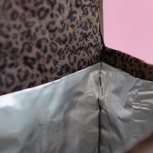 K&ouml;nnte beinhalten: Ge&ouml;ffnete, isolierte Lunchtasche mit braunem und schwarzem Leopardenmuster. Das Innere ist silberfarben. Die Tasche ist gesteppt. Der Hintergrund ist rosa.