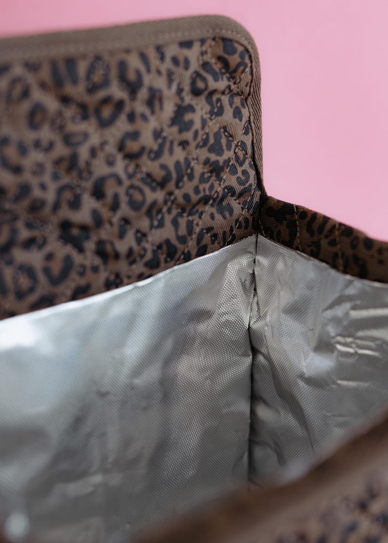 K&ouml;nnte beinhalten: Ge&ouml;ffnete, isolierte Lunchtasche mit braunem und schwarzem Leopardenmuster. Das Innere ist silberfarben. Die Tasche ist gesteppt. Der Hintergrund ist rosa.