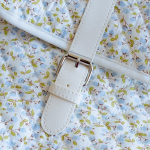 K&ouml;nnte beinhalten: Nahaufnahme eines wei&szlig;en Handtaschenriemens mit silberner Schnalle. Der Riemen ist an einem gesteppten Stoff mit einem Blumenmuster aus blauen Blumen und gr&uuml;nen Bl&auml;ttern auf wei&szlig;em Grund befestigt. Die N&auml;hte sind sichtbar.