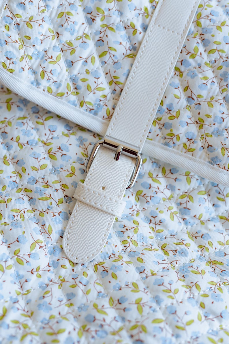 K&ouml;nnte beinhalten: Nahaufnahme eines wei&szlig;en Handtaschenriemens mit silberner Schnalle. Der Riemen ist an einem gesteppten Stoff mit einem Blumenmuster aus blauen Blumen und gr&uuml;nen Bl&auml;ttern auf wei&szlig;em Grund befestigt. Die N&auml;hte sind sichtbar.
