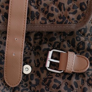 K&ouml;nnte beinhalten: Nahaufnahme eines braun-schwarzen Stoffes mit Leopardenmuster und Steppmuster. Ein hellbrauner Riemen mit silberner Schnalle und Magnetverschluss ist sichtbar. Der Stoff ist dunkelbraun mit schwarzen Flecken.